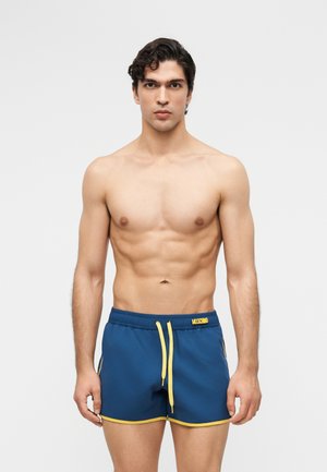 Muskulöser Mann mit kurzen, lockigen Haaren, der blaue Badeshorts mit gelbem Saum und Kordelzug trägt und vor einem schlichten hellen Hintergrund steht.