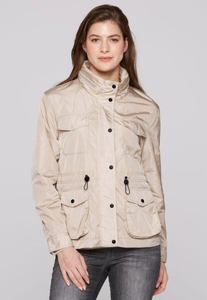 Frau in einer beigen Jacke mit schwarzen Knöpfen und mehreren Fronttaschen, die vor einem schlichten hellgrauen Hintergrund steht.