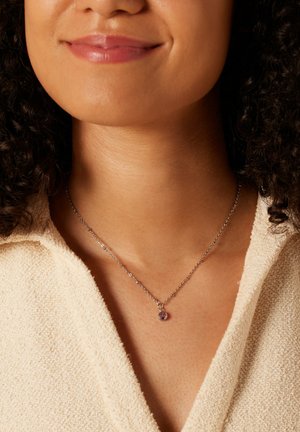 Zilveren halsketting met een delicate ketting en een ronde paarse edelsteen pendant, gedragen tegen een textuur van crèmekleurige stof.