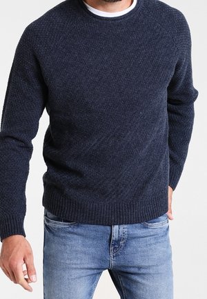 Homme portant un pull en maille texturée bleu marine foncé et un jean denim bleu clair, debout devant un fond blanc uni.