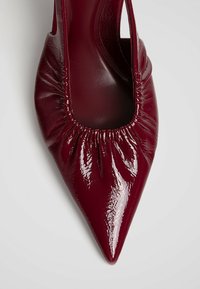 Chaussure plate bordeaux brillante à bout pointu avec un détail élastique froncé autour de l'ouverture, présentée sur un fond clair uni.