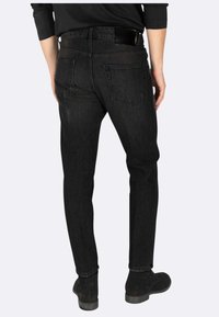 Jeans in denim nero con una vestibilità slim, dotati di due tasche posteriori, un passante in pelle per la cintura e una parte inferiore con risvolto, indossati con stivaletti neri alla caviglia.