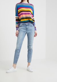 Sweter w biało-multikolorowe paski z dzianiny, zestawiony z jasnoniebieskimi jeansami slim fit z przetarciami oraz białymi trampkami.