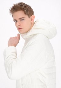 Witte geïsoleerde pufferjas met een gewatteerde textuur en een capuchon. Bevat een ritszak aan de voorkant en verstelbare manchetten. Model kijkt naar links.