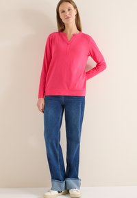 Haut rose à manches longues avec un col henley, associé à un jean bleu évasé avec un ourlet effiloché et des baskets blanches. Tissu doux, coupe décontractée.