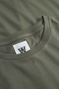 Close-up of olive green t-shirt neckline with a white rectangular tag featuring a black double-A logo.
