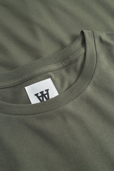 Close-up of olive green t-shirt neckline with a white rectangular tag featuring a black double-A logo.