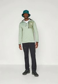 Lichtgroene pullover hoodie met een donkergroen accent, zwarte ritszak, gedragen met zwarte broek en een donkergroene bucket hat; robuuste zwarte schoenen.