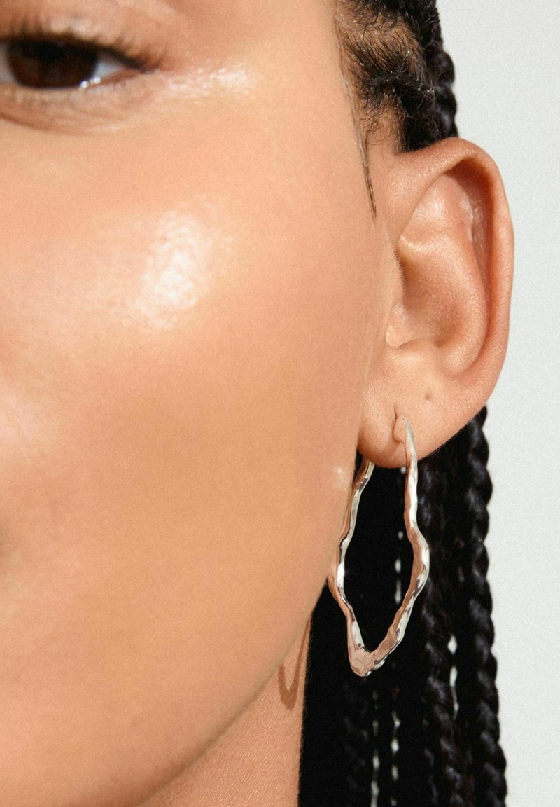 Orecchino a cerchio in argento con un design ondulato, che riflette la luce. Presenta una texture lucida, attaccato a un orecchio forato, con capelli intrecciati visibili.