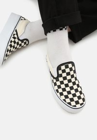 Zapatos slip-on en blanco y negro con diseño de tablero de ajedrez, con parte superior de lona y suela de goma, combinados con calcetines blancos con un ribete negro.