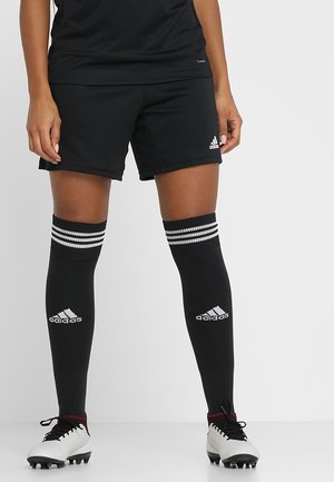 Nogi w czarnych sportowych krótkich spodenkach Adidas, czarne podkolanówki Adidas z białymi paskami oraz białe korki na białej podłodze i tle.