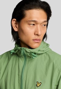 Chaleco con cremallera de color verde claro y capucha, con cordones y un logo negro y amarillo en el pecho. Tejido suave y liviano.