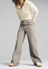 Licht crèmekleurige hoodie met rits en een voorzak, voorzien van een grijs logo, gecombineerd met wijd uitlopende grijze spijkerbroeken. Sneakers met een beige zool.