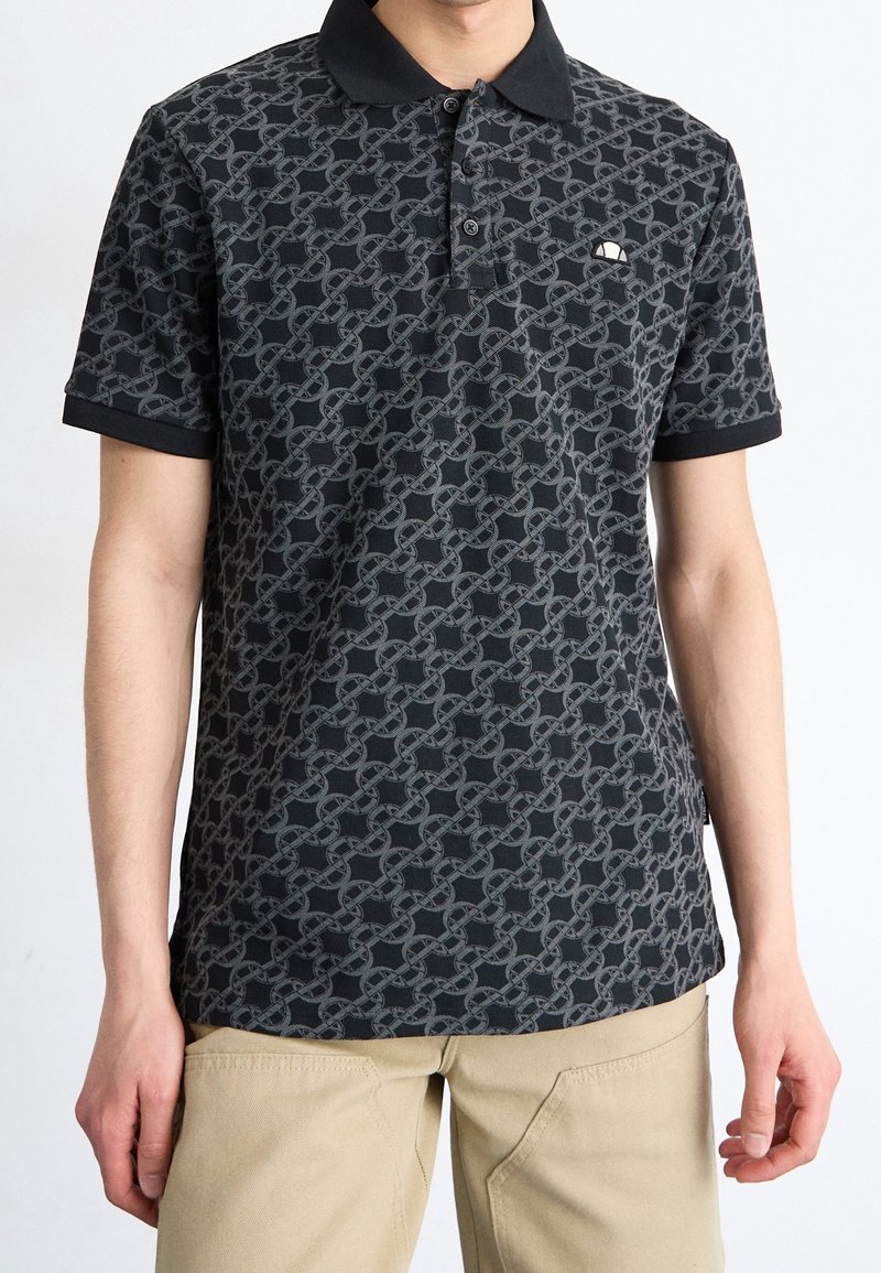 Homme portant un polo noir avec un motif de chaînes entrelacées grises et un petit logo blanc sur la poitrine, pantalon beige, debout devant un fond blanc.