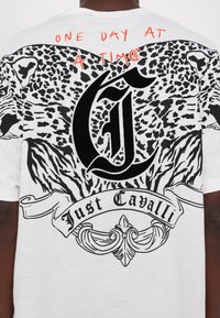 Bílý tričko s krátkým rukávem s černým leopardím potiskem, velkým stylizovaným "G" a textem "Just Cavalli" s červenou ručně psanou frází.