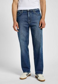 Hombre vestido con jeans rectos azules, zapatillas blancas con rayas negras y una camiseta blanca, de pie contra un fondo liso.