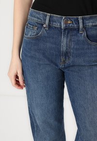 Jean en denim bleu avec une taille haute, design classique à cinq poches et détails de couture jaune. Texture douce avec un léger effet délavé.