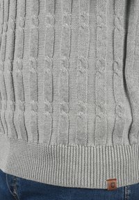 Pull en tricot gris clair avec un motif en câble texturé, un ourlet côtelé et une petite étiquette logo marron sur le côté.