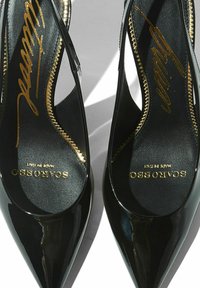 Zapatos de tacón alto de charol negro con puntera afilada y lados recortados. Logo dorado impreso en una plantilla negra. Detalle de costura texturizada.