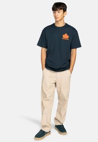 Navy cotton t-shirt with orange leaf logo and text; beige cargo pants with visible stitching; dark teal shoes with textured rubber sole.