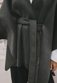 Mörkgrå omslag med ett mjukt, texturerat tyg. Har ett brett bälte och en draperad design, lagd över en vit skjorta och mörka jeans.