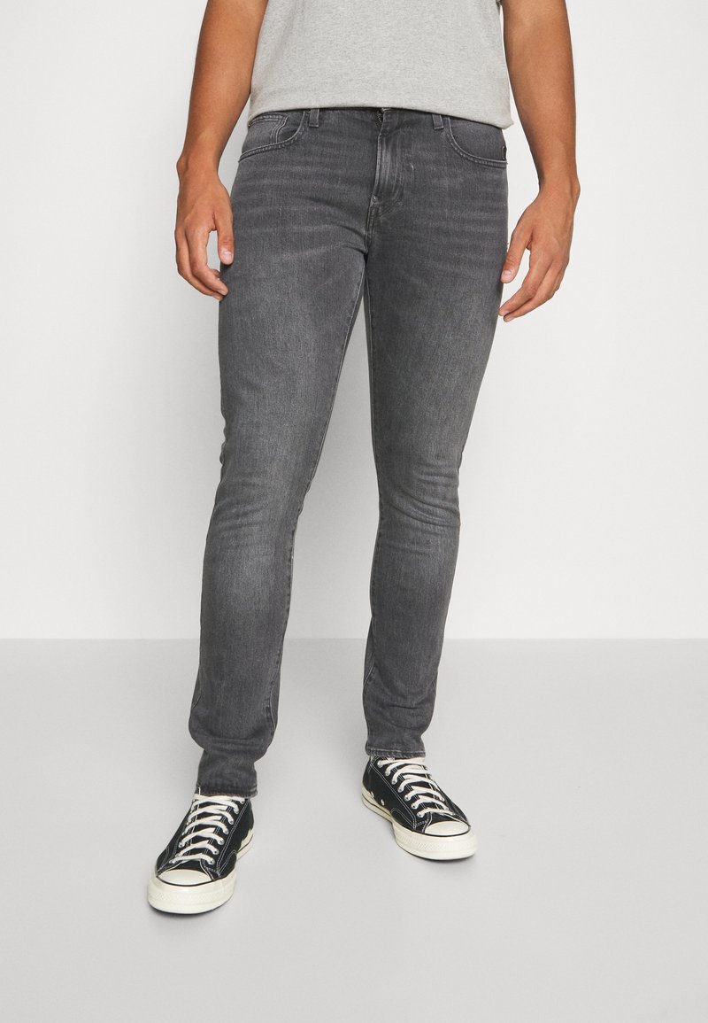 Jeans de mezclilla gris ajustados con cinco bolsillos, un desgaste sutil y costuras en contraste, combinados con zapatillas negras y blancas.