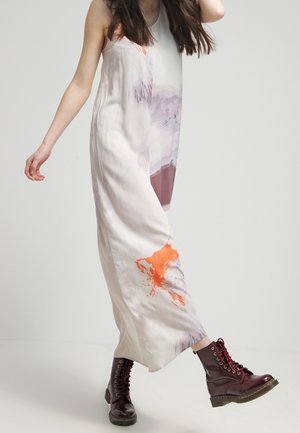 Femme portant une robe blanche sans manches, longue jusqu'au sol, avec des motifs abstraits orange et violet, associée à des bottes lacées rouge foncé.