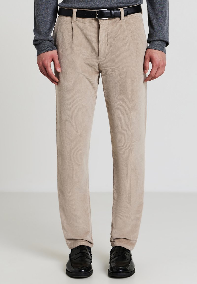 Uomo in pantaloni corduroy beige, cintura nera, maglione grigio e mocassini in pelle nera, in piedi davanti a uno sfondo semplice.