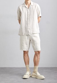Camicia button-up beige in lino a maniche corte abbinata a pantaloni corti dello stesso colore. Indossata con scarpe slip-on beige e calzini beige chiaro.