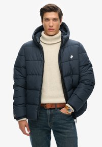 Marineblå pufferjakke med vandret quiltet mønster, hætte, lynlåslukning og lille logoaccent. Båret over en cremefarvet turtleneck og denim.