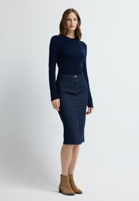 Laivastonsininen pitkähihainen paita yhdistettynä polvenpituiseen teksturoituun hameeseen. Vyötäröllä nappiyksityiskohta ja ruskeat mokkanahkaiset nilkkurit.