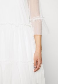 Robe blanche présentant un design à étages avec des bords volantés, des manches courtes en tissu transparent et un tissu texturé, offrant une apparence légère.