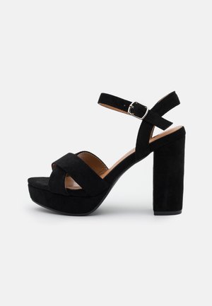 Sort ruskind sandal med plateau, tykt hæl og ankelrem, med et krydsrem-design foran og en tekstureret overflade.