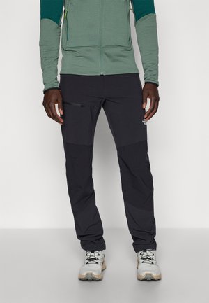 Mann trägt schwarze Outdoorhose mit Reißverschlusstasche und graue Wanderschuhe, steht vor einem einfarbigen hellen Hintergrund.