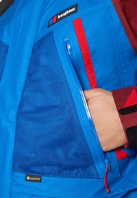 Mėlyna nepralaidi vandeniui striukė su tekstūruotu paviršiumi, turinti raudonus akcentus, užtrauktuku užsidarančią kišenę ir GORE-TEX etiketę apatinėje panelėje.