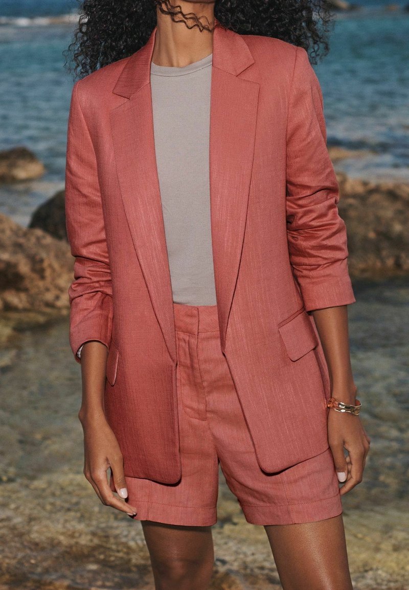 Blazer en lin rose avec short assorti, présentant un design structuré, des poches plaquées et une finition texturée, assorti à un haut gris.