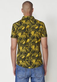 Cămașă cu mâneci scurte, cu un imprimeu de frunze de palmier galbene și negre, având un guler standard și o margine dreaptă. Purtată cu blugi denim blue.