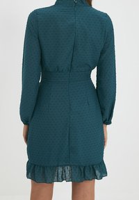 Teele Kleid mit langen Ärmeln, strukturiertem Punktemuster, tailliertem Schnitt und Rüschensaum. Reißverschluss hinten und hoher Kragen.