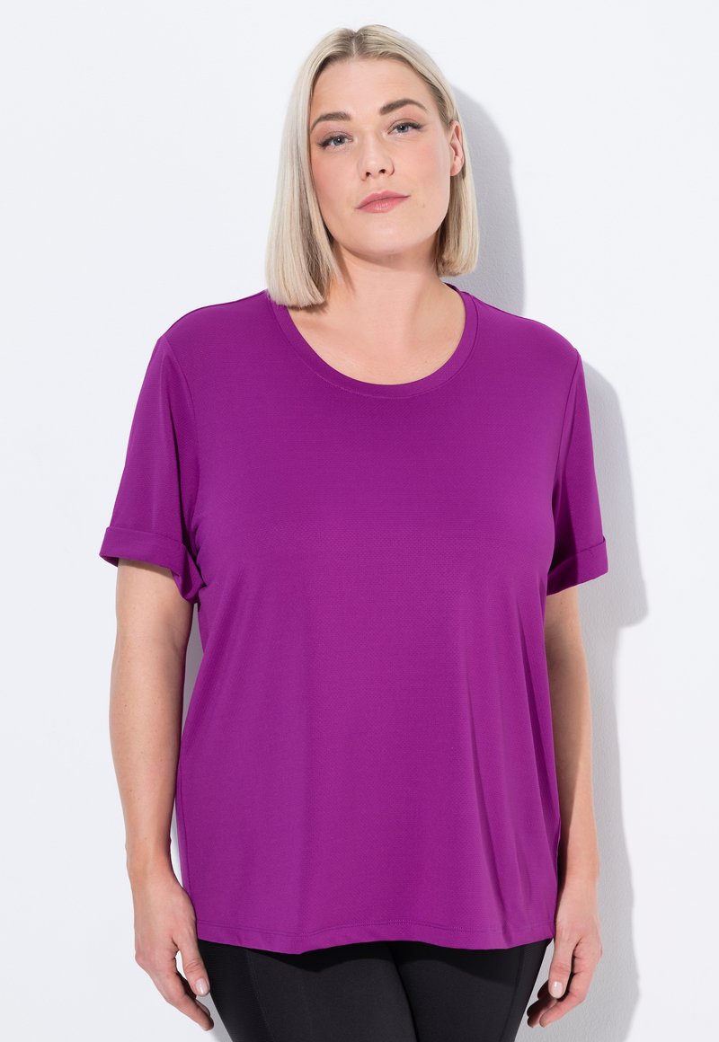 Lyshåret kvinde iført en lilla kortærmet skjorte og sorte bukser, som står opad en hvid væg, vender fremad med et neutralt udtryk.