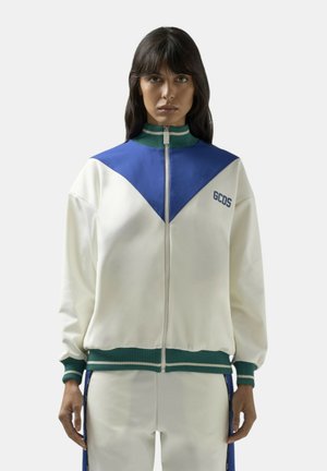 Donna che indossa una giacca bianca con zip con dettaglio triangolare blu sul petto, colletto e polsini verdi a coste, logo "GCDS" sul petto a sinistra e pantaloni coordinati.