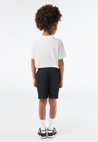 Enfant debout, dos à la caméra, portant un t-shirt blanc, un short noir, des chaussettes blanches et des baskets noires sur un fond uni.