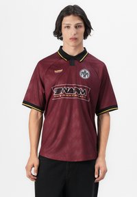 Jonge man met halflang donker haar draagt een bordeaux polo met zwarte kraag en gele bies, met "SWARM"- en embleemlogo's.