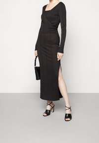 Robe maxi noire à côtes avec des manches longues, un décolleté carré et une fente sur le côté, associée à des sandales à talons noires et un petit sac à main noir.
