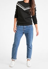 Sudadera negra con rayas en forma de chevron blancas, vaqueros de denim azul y deportivas blancas. Ajuste relajado, puños enrollados en los vaqueros, texturas suaves.