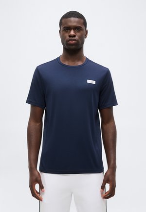 Giovane uomo con t-shirt sportiva a maniche corte blu navy e pantaloncini bianchi, in piedi davanti a uno sfondo chiaro e uniforme.