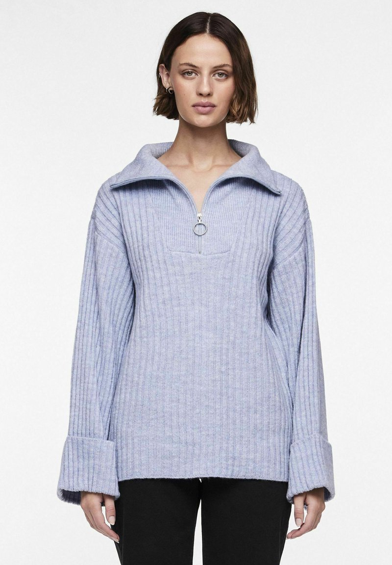 Šviesiai mėlynas įstrižas megztinis su plačiu atlenkiamu apykakliu, pusiau užtrauktuku priekyje ir per dideliais rankogaliais. Minkšta tekstūra ir laisvas kirpimas.