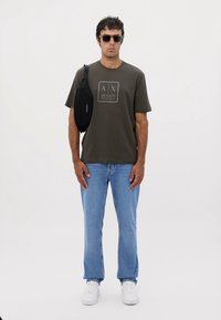 Camiseta de algodón verde oscuro con un logo central, jeans azul claro, zapatillas blancas y una bolsa cruzada negra, llevada por un modelo masculino.