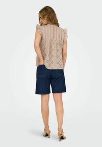 Hellbeige Bluse aus Broderie Anglaise mit Rüschensleeves, kombiniert mit dunklen Denim-Shorts, die Taschen und einen lockeren Schnitt präsentieren. Neutrale Schuhe runden das Outfit ab.