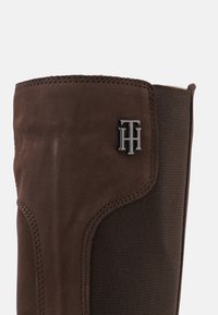 Brązowy zamszowy but z elastycznymi panelami, z teksturowaną powierzchnią i metalowym akcentem logo. Okrągły kształt z wzmocnionymi detalami szycia.