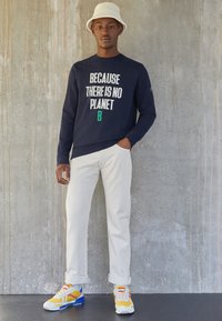 Navy sweatshirt with "BECAUSE THERE IS NO PLANET B" in white and green text, paired with white pants and colorful sneakers.